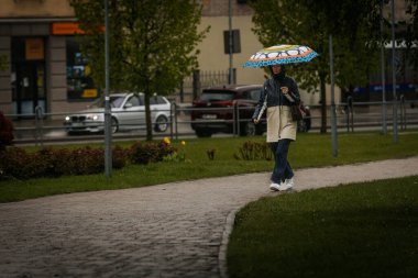 İki tonluk yağmurluk ve kot pantolon giymiş bir kadın, elinde canlı, desenli bir şemsiye, arka planda yeşil ve kentsel binalarla ıslak tuğla bir yolda yürüyor..