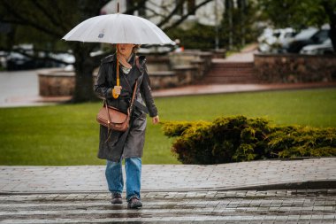 İki tonluk yağmurluk ve kot pantolon giymiş bir kadın, elinde canlı, desenli bir şemsiye, arka planda yeşil ve kentsel binalarla ıslak tuğla bir yolda yürüyor..