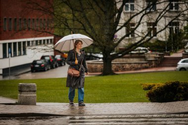 İki tonluk yağmurluk ve kot pantolon giymiş bir kadın, elinde canlı, desenli bir şemsiye, arka planda yeşil ve kentsel binalarla ıslak tuğla bir yolda yürüyor..