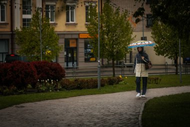 İki tonluk yağmurluk ve kot pantolon giymiş bir kadın, elinde canlı, desenli bir şemsiye, arka planda yeşil ve kentsel binalarla ıslak tuğla bir yolda yürüyor..