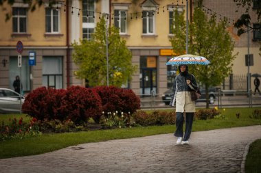 İki tonluk yağmurluk ve kot pantolon giymiş bir kadın, elinde canlı, desenli bir şemsiye, arka planda yeşil ve kentsel binalarla ıslak tuğla bir yolda yürüyor..
