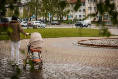 İki tonluk yağmurluk ve kot pantolon giymiş bir kadın, elinde canlı, desenli bir şemsiye, arka planda yeşil ve kentsel binalarla ıslak tuğla bir yolda yürüyor..