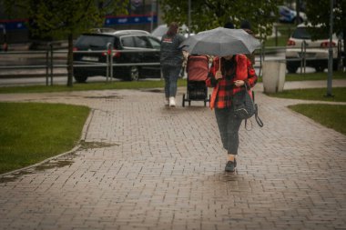 İki tonluk yağmurluk ve kot pantolon giymiş bir kadın, elinde canlı, desenli bir şemsiye, arka planda yeşil ve kentsel binalarla ıslak tuğla bir yolda yürüyor..
