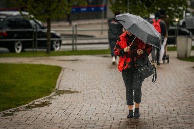 İki tonluk yağmurluk ve kot pantolon giymiş bir kadın, elinde canlı, desenli bir şemsiye, arka planda yeşil ve kentsel binalarla ıslak tuğla bir yolda yürüyor..