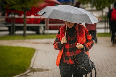 İki tonluk yağmurluk ve kot pantolon giymiş bir kadın, elinde canlı, desenli bir şemsiye, arka planda yeşil ve kentsel binalarla ıslak tuğla bir yolda yürüyor..