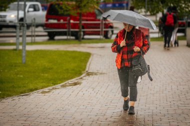 İki tonluk yağmurluk ve kot pantolon giymiş bir kadın, elinde canlı, desenli bir şemsiye, arka planda yeşil ve kentsel binalarla ıslak tuğla bir yolda yürüyor..