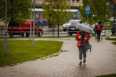 İki tonluk yağmurluk ve kot pantolon giymiş bir kadın, elinde canlı, desenli bir şemsiye, arka planda yeşil ve kentsel binalarla ıslak tuğla bir yolda yürüyor..