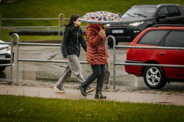 İki tonluk yağmurluk ve kot pantolon giymiş bir kadın, elinde canlı, desenli bir şemsiye, arka planda yeşil ve kentsel binalarla ıslak tuğla bir yolda yürüyor..