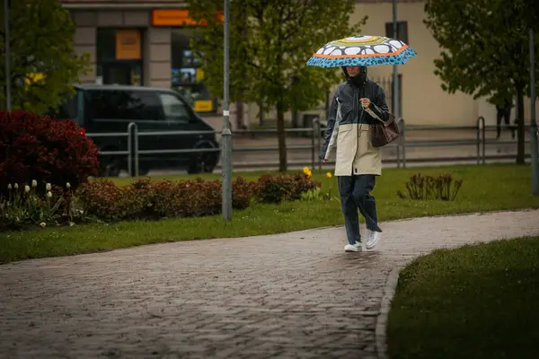 İki tonluk yağmurluk ve kot pantolon giymiş bir kadın, elinde canlı, desenli bir şemsiye, arka planda yeşil ve kentsel binalarla ıslak tuğla bir yolda yürüyor..