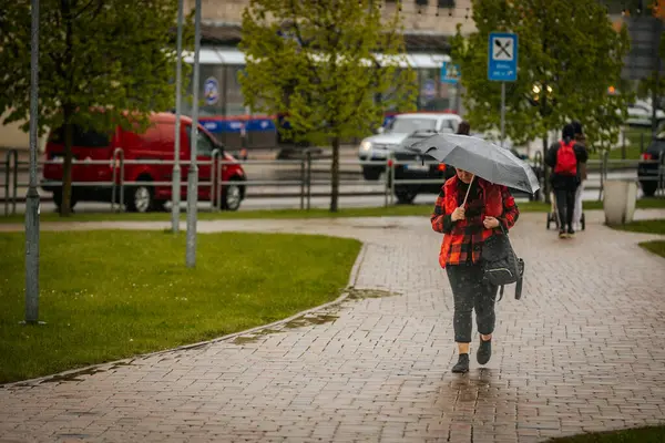 İki tonluk yağmurluk ve kot pantolon giymiş bir kadın, elinde canlı, desenli bir şemsiye, arka planda yeşil ve kentsel binalarla ıslak tuğla bir yolda yürüyor..