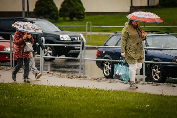 İki tonluk yağmurluk ve kot pantolon giymiş bir kadın, elinde canlı, desenli bir şemsiye, arka planda yeşil ve kentsel binalarla ıslak tuğla bir yolda yürüyor..