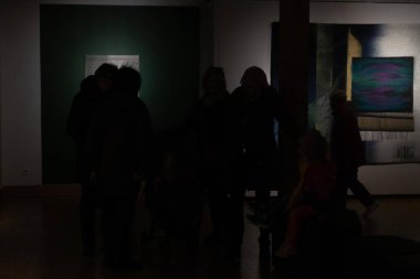 Çocuklar da dahil olmak üzere bir grup insan, hafif ışıklandırılmış bir galeride aydınlatılmış tekstil sanat eserlerine bakıyorlar. Gölgeler ve kontrastlar derin bir atmosfer ekler. 17.05.2025 Valmiera, Letonya