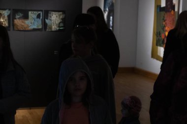 Çocuklar da dahil olmak üzere bir grup insan, hafif ışıklandırılmış bir galeride aydınlatılmış tekstil sanat eserlerine bakıyorlar. Gölgeler ve kontrastlar derin bir atmosfer ekler. 17.05.2025 Valmiera, Letonya