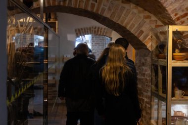 Bir grup insan müze gibi bir yerde toplanıyor ve duvarlarda ve tavanda geleneksel ahşap aletler sergileniyor. Işıklandırma sıcak ve bastırılmış. 17.05.2025 Valmiera, Letonya