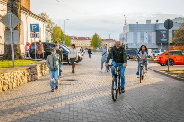 Neşeli bir adam ve kadın bisiklet sürüyorlar, arka planda yayalar ve arabalarla, güneşli bir günde gülümsüyorlar ve eğleniyorlar. 17.05.2025 Valmiera, Letonya