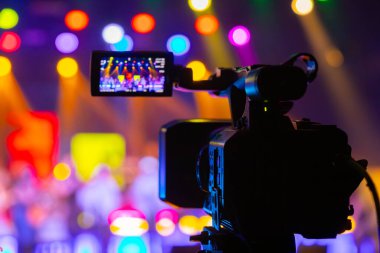 Profesyonel video kamera canlı bir konser ya da etkinlik kaydediyor, sahneyi gösteren bir ekran ve arka planda parlak renkli ışıklar. 22.05.2025 Valmiera, Letonya