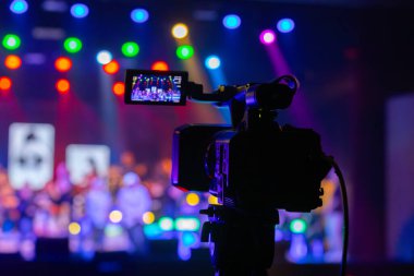 Profesyonel video kamera canlı bir konser ya da etkinlik kaydediyor, sahneyi gösteren bir ekran ve arka planda parlak renkli ışıklar. 22.05.2025 Valmiera, Letonya