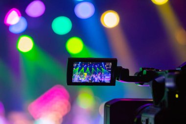 Profesyonel video kamera canlı bir konser ya da etkinlik kaydediyor, sahneyi gösteren bir ekran ve arka planda parlak renkli ışıklar. 22.05.2025 Valmiera, Letonya