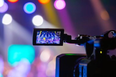 Profesyonel video kamera canlı bir konser ya da etkinlik kaydediyor, sahneyi gösteren bir ekran ve arka planda parlak renkli ışıklar. 22.05.2025 Valmiera, Letonya