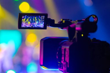 Profesyonel video kamera canlı bir konser ya da etkinlik kaydediyor, sahneyi gösteren bir ekran ve arka planda parlak renkli ışıklar. 22.05.2025 Valmiera, Letonya