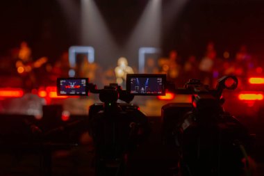 Parlak spot ışıkları ve canlı bokeh ile aydınlatılmış bir konser sahnesini kaydeden iki video kamera, canlı bir yapım düzeneği sergiliyor. 22.05.2025 Valmiera, Letonya