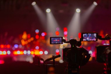 Parlak spot ışıkları ve canlı bokeh ile aydınlatılmış bir konser sahnesini kaydeden iki video kamera, canlı bir yapım düzeneği sergiliyor. 22.05.2025 Valmiera, Letonya