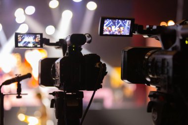 Parlak spot ışıkları ve canlı bokeh ile aydınlatılmış bir konser sahnesini kaydeden iki video kamera, canlı bir yapım düzeneği sergiliyor. 22.05.2025 Valmiera, Letonya