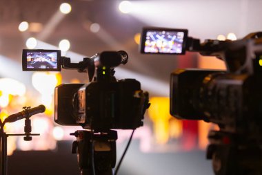 Parlak spot ışıkları ve canlı bokeh ile aydınlatılmış bir konser sahnesini kaydeden iki video kamera, canlı bir yapım düzeneği sergiliyor. 22.05.2025 Valmiera, Letonya
