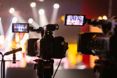 Parlak spot ışıkları ve canlı bokeh ile aydınlatılmış bir konser sahnesini kaydeden iki video kamera, canlı bir yapım düzeneği sergiliyor. 22.05.2025 Valmiera, Letonya