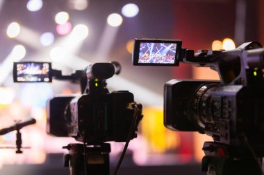 Parlak spot ışıkları ve canlı bokeh ile aydınlatılmış bir konser sahnesini kaydeden iki video kamera, canlı bir yapım düzeneği sergiliyor. 22.05.2025 Valmiera, Letonya
