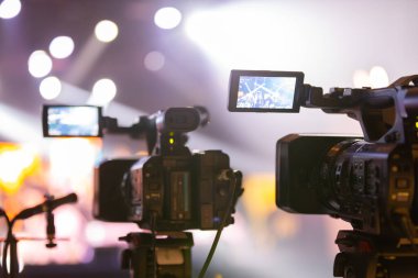 Parlak spot ışıkları ve canlı bokeh ile aydınlatılmış bir konser sahnesini kaydeden iki video kamera, canlı bir yapım düzeneği sergiliyor. 22.05.2025 Valmiera, Letonya