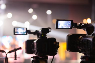 Parlak spot ışıkları ve canlı bokeh ile aydınlatılmış bir konser sahnesini kaydeden iki video kamera, canlı bir yapım düzeneği sergiliyor. 22.05.2025 Valmiera, Letonya