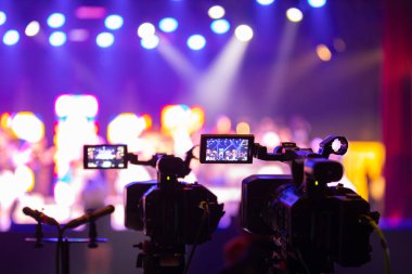 Parlak spot ışıkları ve canlı bokeh ile aydınlatılmış bir konser sahnesini kaydeden iki video kamera, canlı bir yapım düzeneği sergiliyor. 22.05.2025 Valmiera, Letonya