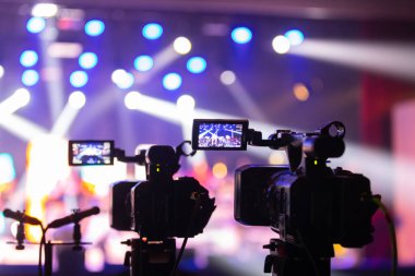 Parlak spot ışıkları ve canlı bokeh ile aydınlatılmış bir konser sahnesini kaydeden iki video kamera, canlı bir yapım düzeneği sergiliyor. 22.05.2025 Valmiera, Letonya