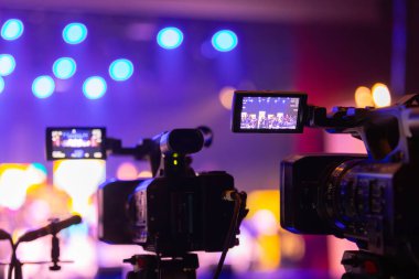 Parlak spot ışıkları ve canlı bokeh ile aydınlatılmış bir konser sahnesini kaydeden iki video kamera, canlı bir yapım düzeneği sergiliyor. 22.05.2025 Valmiera, Letonya