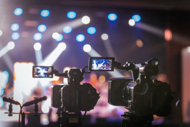 Parlak spot ışıkları ve canlı bokeh ile aydınlatılmış bir konser sahnesini kaydeden iki video kamera, canlı bir yapım düzeneği sergiliyor. 22.05.2025 Valmiera, Letonya
