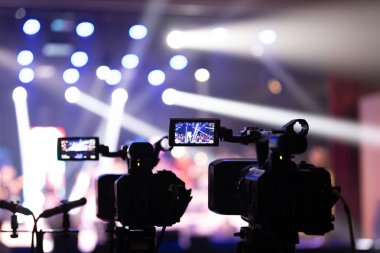 Parlak spot ışıkları ve canlı bokeh ile aydınlatılmış bir konser sahnesini kaydeden iki video kamera, canlı bir yapım düzeneği sergiliyor. 22.05.2025 Valmiera, Letonya