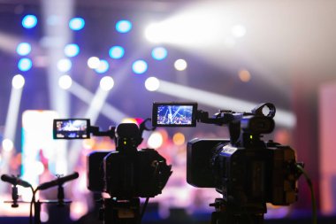 Parlak spot ışıkları ve canlı bokeh ile aydınlatılmış bir konser sahnesini kaydeden iki video kamera, canlı bir yapım düzeneği sergiliyor. 22.05.2025 Valmiera, Letonya