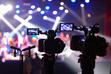 Parlak spot ışıkları ve canlı bokeh ile aydınlatılmış bir konser sahnesini kaydeden iki video kamera, canlı bir yapım düzeneği sergiliyor. 22.05.2025 Valmiera, Letonya