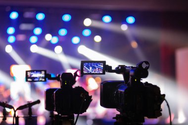 Parlak spot ışıkları ve canlı bokeh ile aydınlatılmış bir konser sahnesini kaydeden iki video kamera, canlı bir yapım düzeneği sergiliyor. 22.05.2025 Valmiera, Letonya