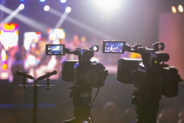 Parlak spot ışıkları ve canlı bokeh ile aydınlatılmış bir konser sahnesini kaydeden iki video kamera, canlı bir yapım düzeneği sergiliyor. 22.05.2025 Valmiera, Letonya
