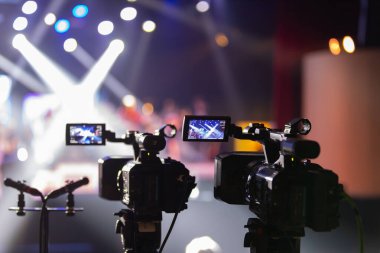 Parlak spot ışıkları ve canlı bokeh ile aydınlatılmış bir konser sahnesini kaydeden iki video kamera, canlı bir yapım düzeneği sergiliyor. 22.05.2025 Valmiera, Letonya