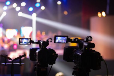 Parlak spot ışıkları ve canlı bokeh ile aydınlatılmış bir konser sahnesini kaydeden iki video kamera, canlı bir yapım düzeneği sergiliyor. 22.05.2025 Valmiera, Letonya