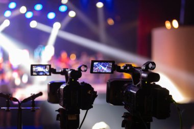 Parlak spot ışıkları ve canlı bokeh ile aydınlatılmış bir konser sahnesini kaydeden iki video kamera, canlı bir yapım düzeneği sergiliyor. 22.05.2025 Valmiera, Letonya