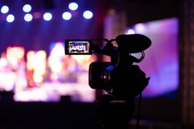 Profesyonel video kamera canlı bir konser ya da etkinlik kaydediyor, sahneyi gösteren bir ekran ve arka planda parlak renkli ışıklar. 22.05.2025 Valmiera, Letonya