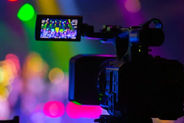 Profesyonel video kamera canlı bir konser ya da etkinlik kaydediyor, sahneyi gösteren bir ekran ve arka planda parlak renkli ışıklar. 22.05.2025 Valmiera, Letonya