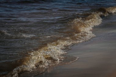 Güneş ışığı altında kıyıya yaklaşan küçük köpüklü bir dalganın yakın çekimi, koyu mavi sudaki dalgalanmaları ve dokuları vurguluyor..