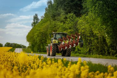 Kırmızı saban ekipmanlı büyük yeşil bir traktör, mavi gökyüzünün altında çiçek açan sarı çiçekler ve yemyeşil ağaçlarla dolu kırsal bir yolda ilerliyor..