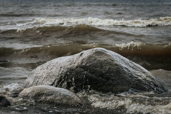 Темная скала, частично погруженная в морскую воду с маленькими волнами, разбивающимися на песчаном пляже под солнечным светом.