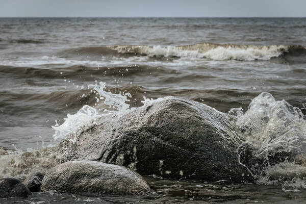 Темная скала, частично погруженная в морскую воду с маленькими волнами, разбивающимися на песчаном пляже под солнечным светом.